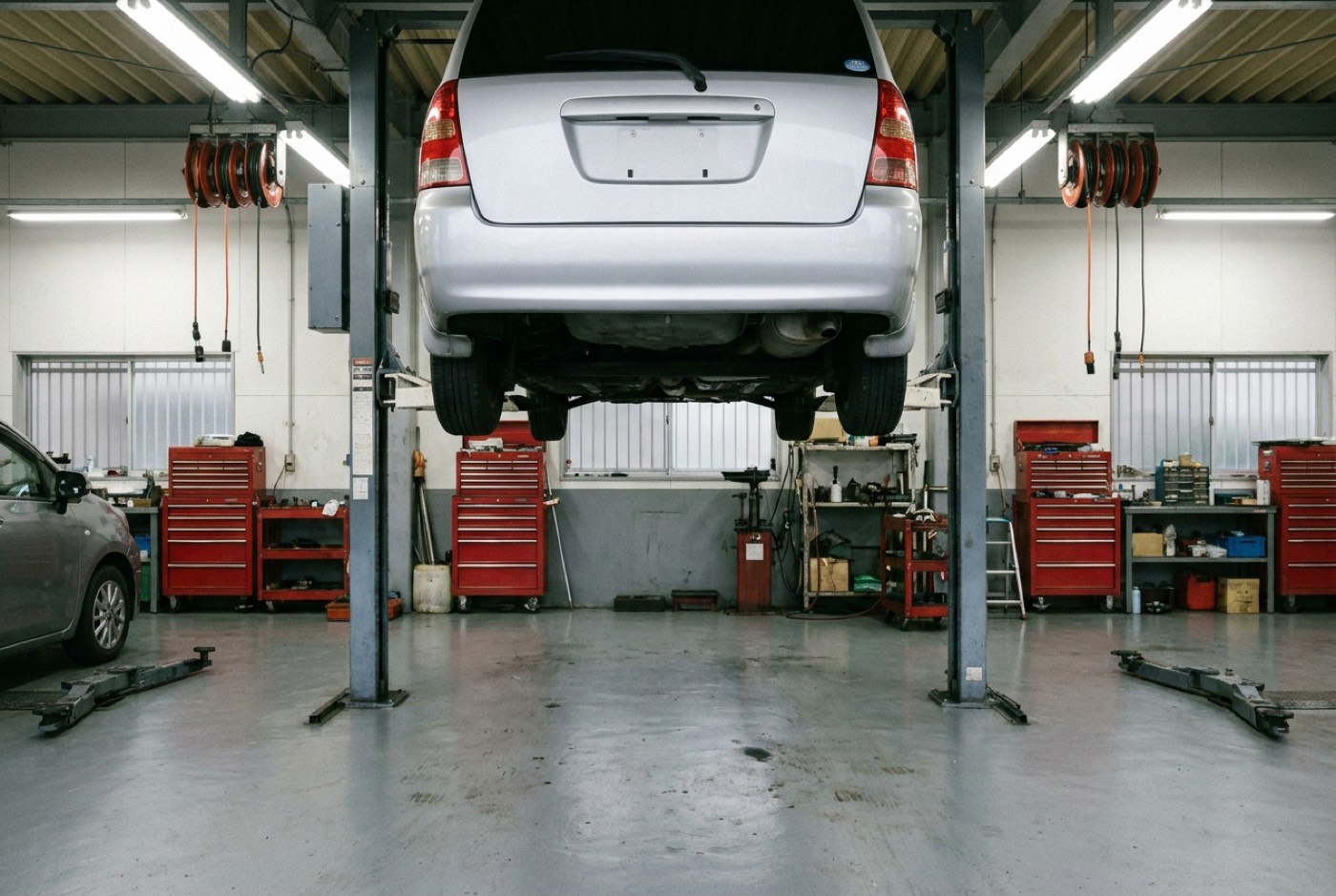 蛍光灯で明るく照らされた整備工場の作業ベイ。2柱リフトに乗った1台の乗用車と、壁際に並ぶ赤い工具キャビネット