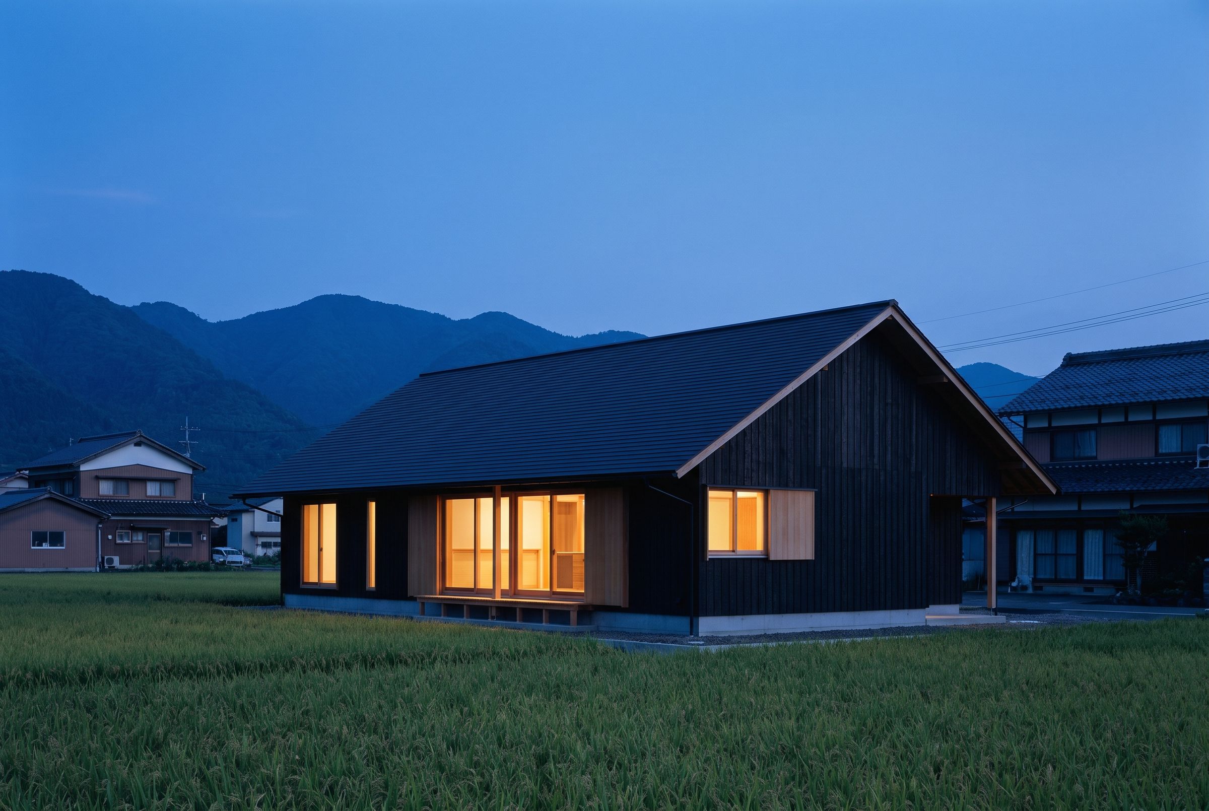 夕暮れの田んぼの向こうに建つ、黒い杉板張りの切妻屋根の平屋。大きな窓から明かりがこぼれる