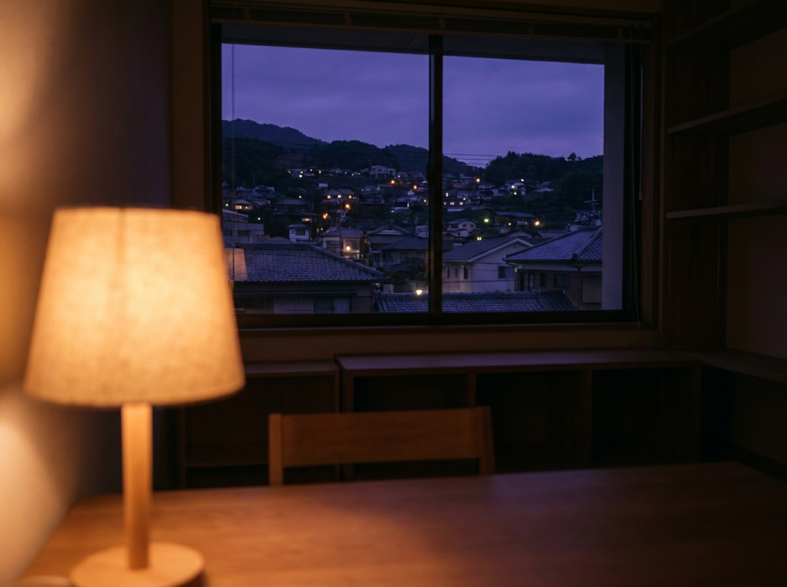 夜の窓辺。手前にテーブルランプ、窓の外に小さな町の灯りが散っている
