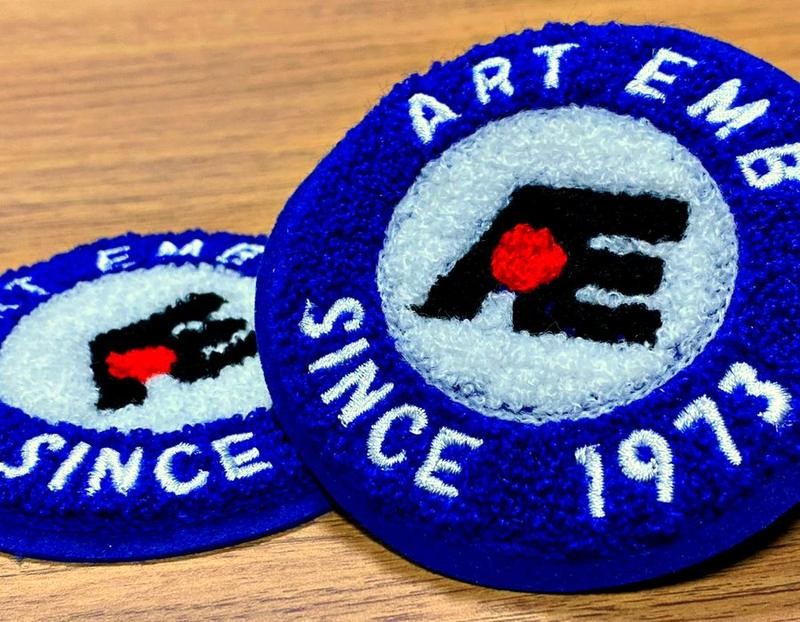 ART EMB SINCE 1973 と刺繍された、青と白の丸いワッペン2枚を木の机に置いた写真